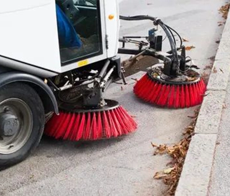 The red brushes of a street sweeper sweep up leaves along a sidewalk.