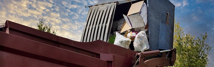 A garbage truck dumps refuse into a large maroon dumpster.