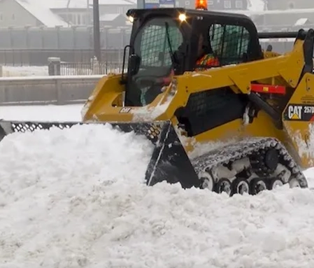 A yellow Cat snow plow pushes through a snow-filled parking lot.