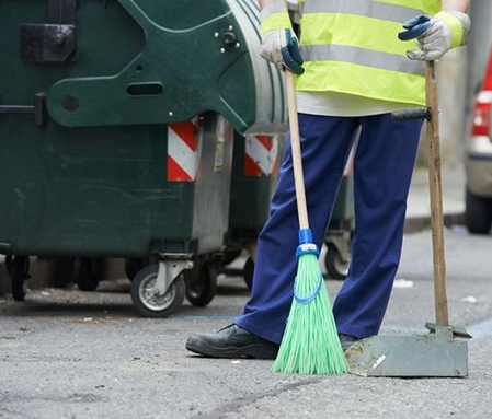 An employee of Naco Commercial Propety Solutions wears blue pants and a yellow high visibility jacket. They hold a broom in one hand and a dustpan in the other while sweeping a parking lot.