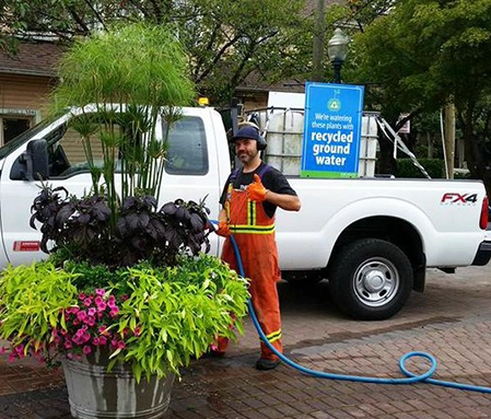 A Naco Commercial Property Solutions team member wears orange high visibility coveralls and gives a thumbs up while watering plants in front of a white pickup truck.