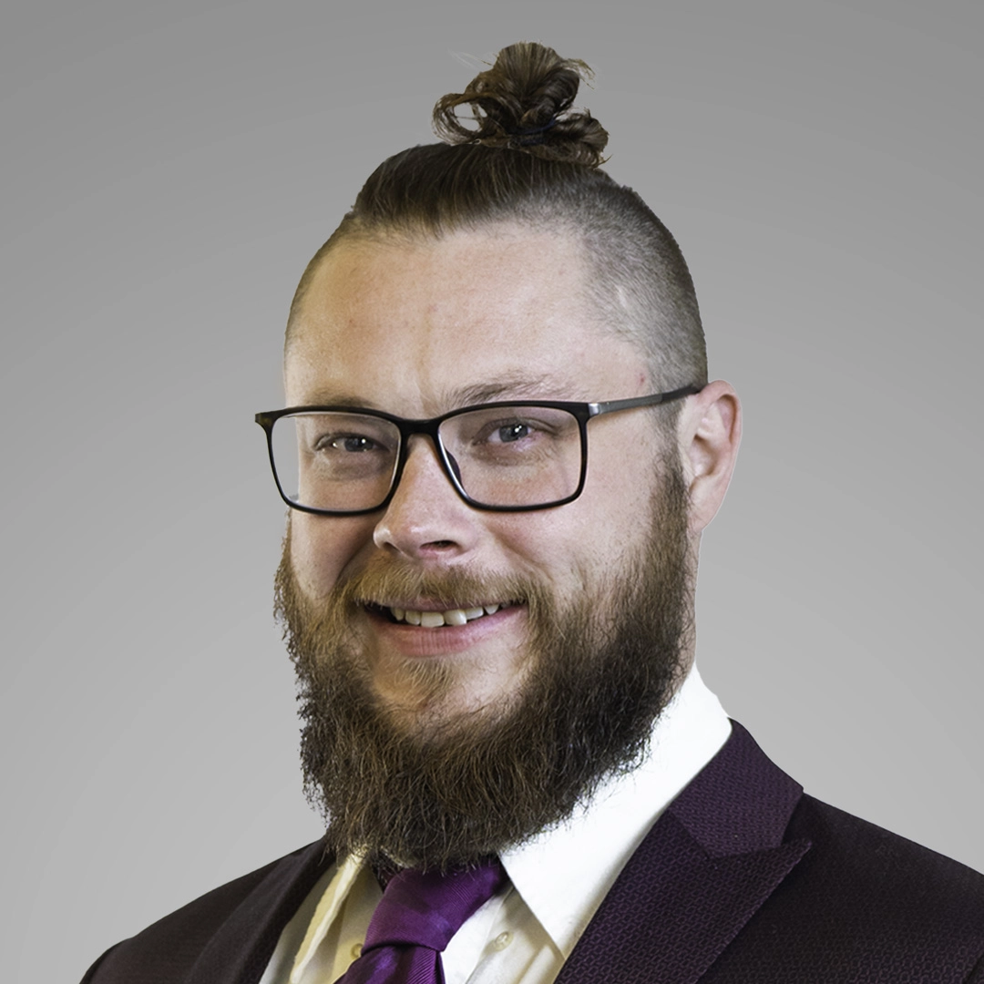 Eric Laffoley, the Project Manager and Lead Estimator at Naco Commercial Property Solutions smiles at the camera. He wears a deep purple suit, purple tie, white shirt, and black glasses.