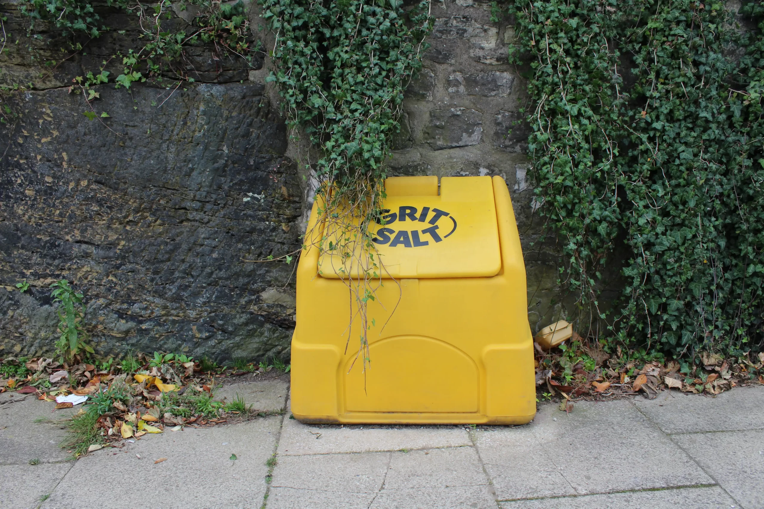 A yellow grit bin on a side walk in front of a rock wall, with green vines hanging around it.